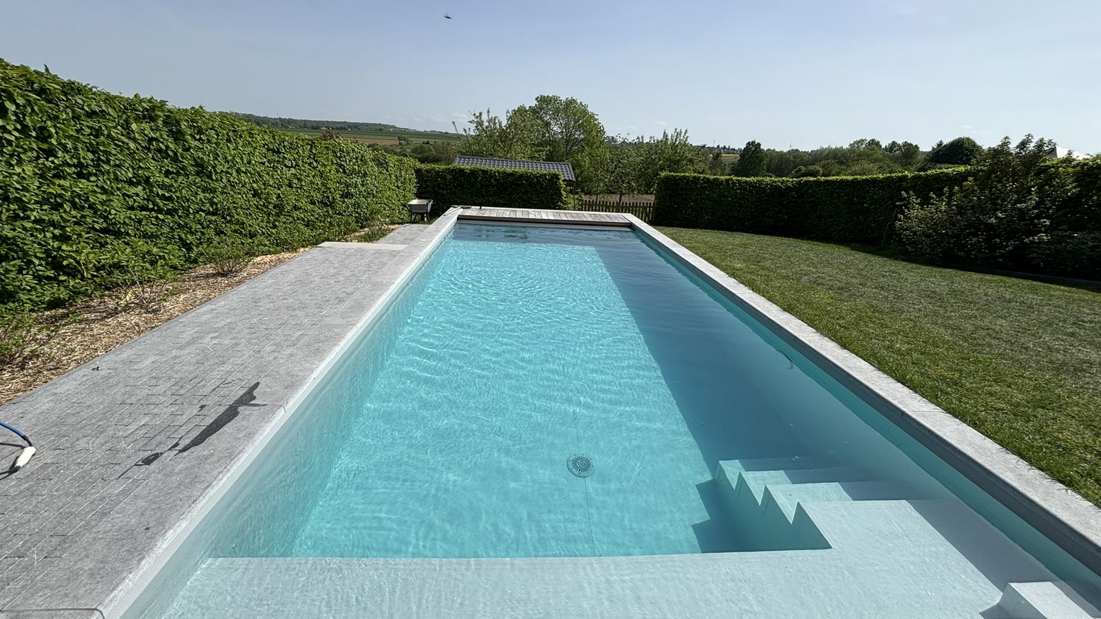 Longue piscine enterrée à l'eau turquoise, entourée de pelouse et de haies
